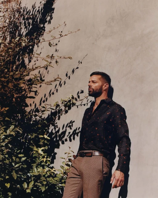 Adult man in patterned shirt poses against wall with plants. He wears a belt with brown trousers and a serious expression.