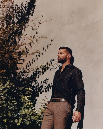 Adult man in patterned shirt poses against wall with plants. He wears a belt with brown trousers and a serious expression.