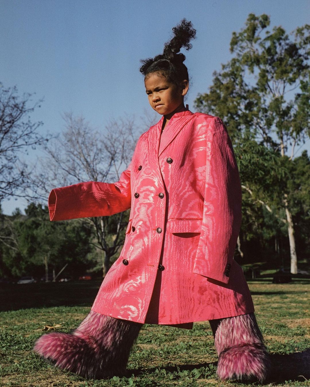 Young girl in a pink patterned coat and furry leg coverings walking in a grassy field with trees, beneath a blue sky.