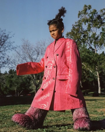 Young girl in a pink patterned coat and furry leg coverings walking in a grassy field with trees, beneath a blue sky.
