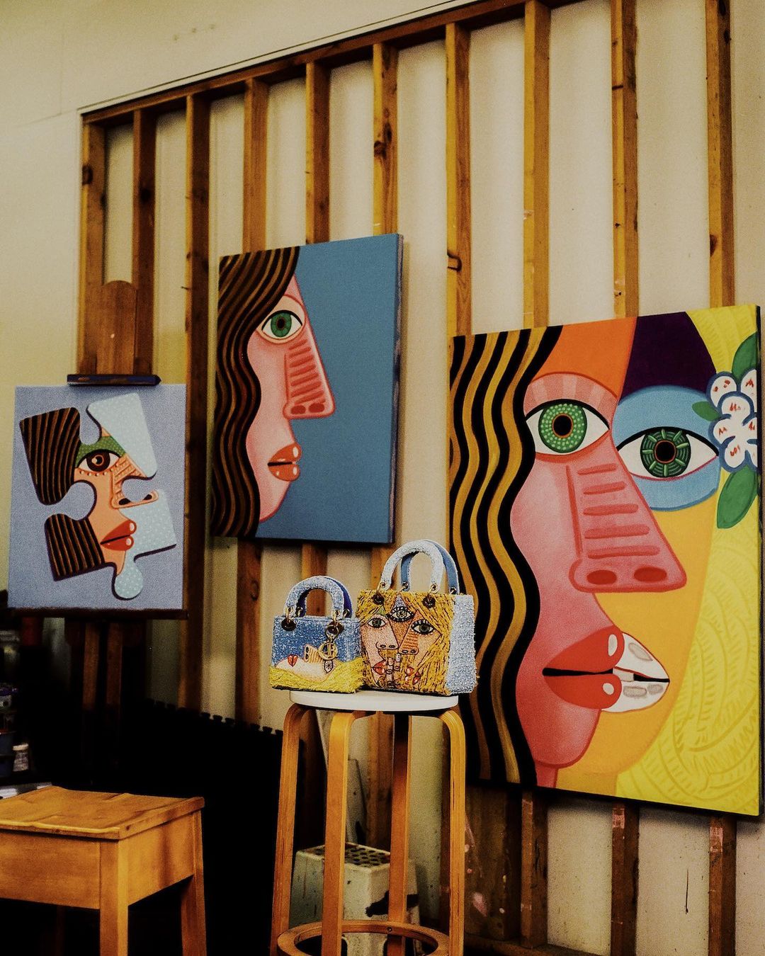 Three face-themed artworks displayed in an art studio, complemented by two embellished handbags on a wooden stool, all against a wooden wall.