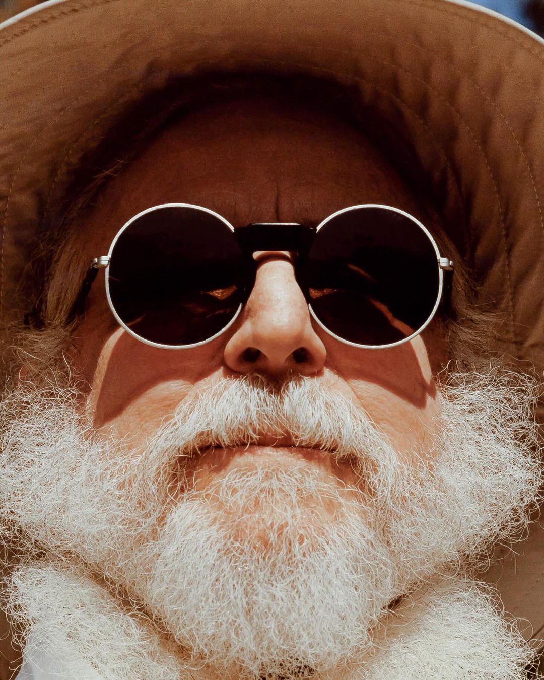 Portrait of an adult wearing a safari hat and round sunglasses. The adult has a long white beard and fair skin. The background is indistinct.