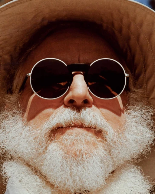 Portrait of an adult wearing a safari hat and round sunglasses. The adult has a long white beard and fair skin. The background is indistinct.