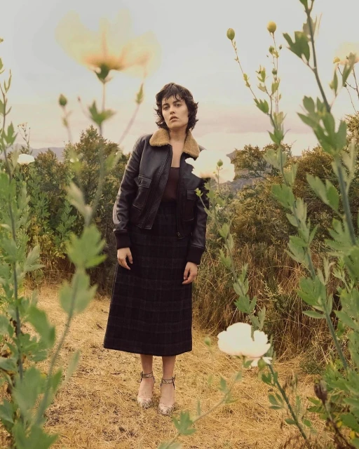 Woman in vintage-style bomber jacket and skirt stands in a field of white flowers with distant hills under soft, neutral lighting.