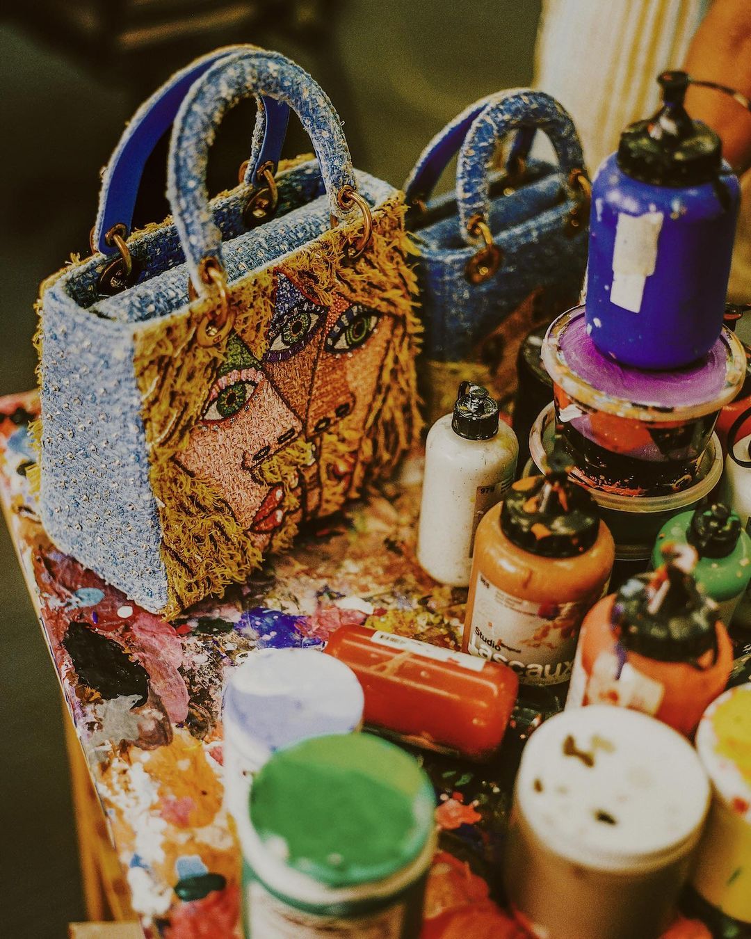 Close-up of two miniature denim handbags, embellished with artistic facial designs, displayed on a table covered with colorful paint supplies.