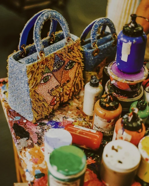 Close-up of two miniature denim handbags, embellished with artistic facial designs, displayed on a table covered with colorful paint supplies.