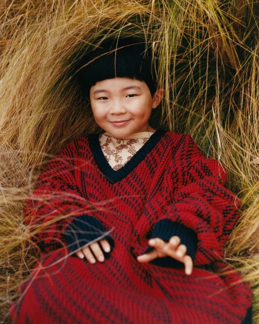 Young child with black hair smiles while lying in dry grass, wearing a red knit sweater in an eye-level portrait.