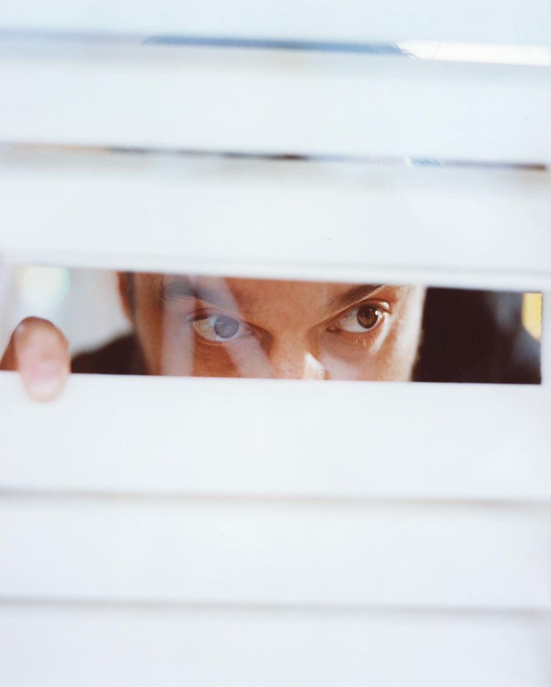 Eye-level view of an adult male peering through horizontal white blinds with an inquisitive expression, conveying a sense of mystery.