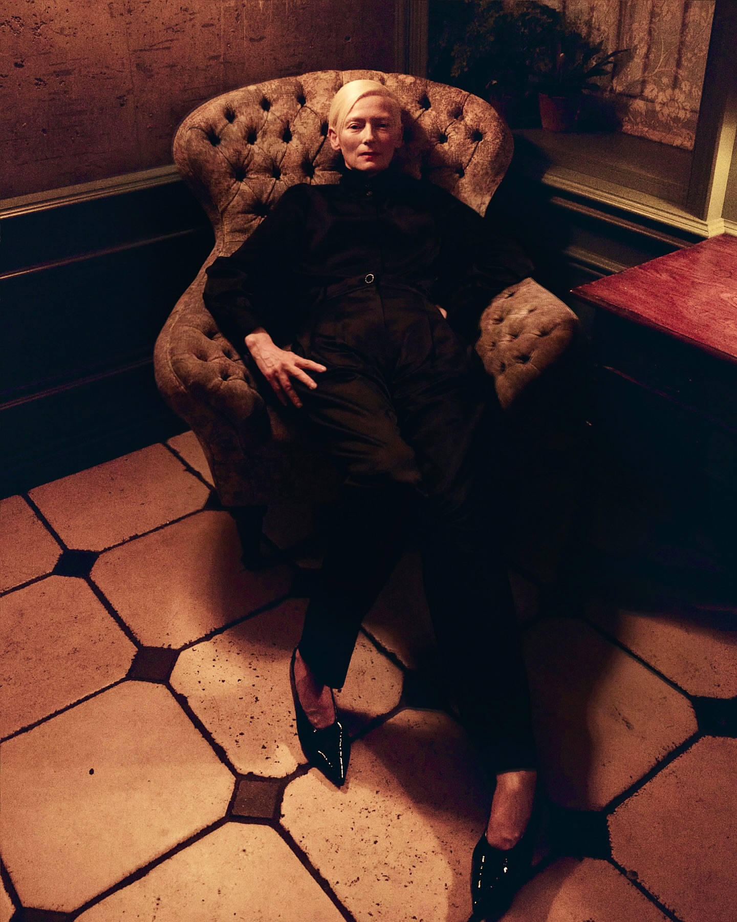 A fair-skinned adult woman with blond hair sits in a tufted armchair against a dark, textured wall on a tiled floor, wearing dark clothing.