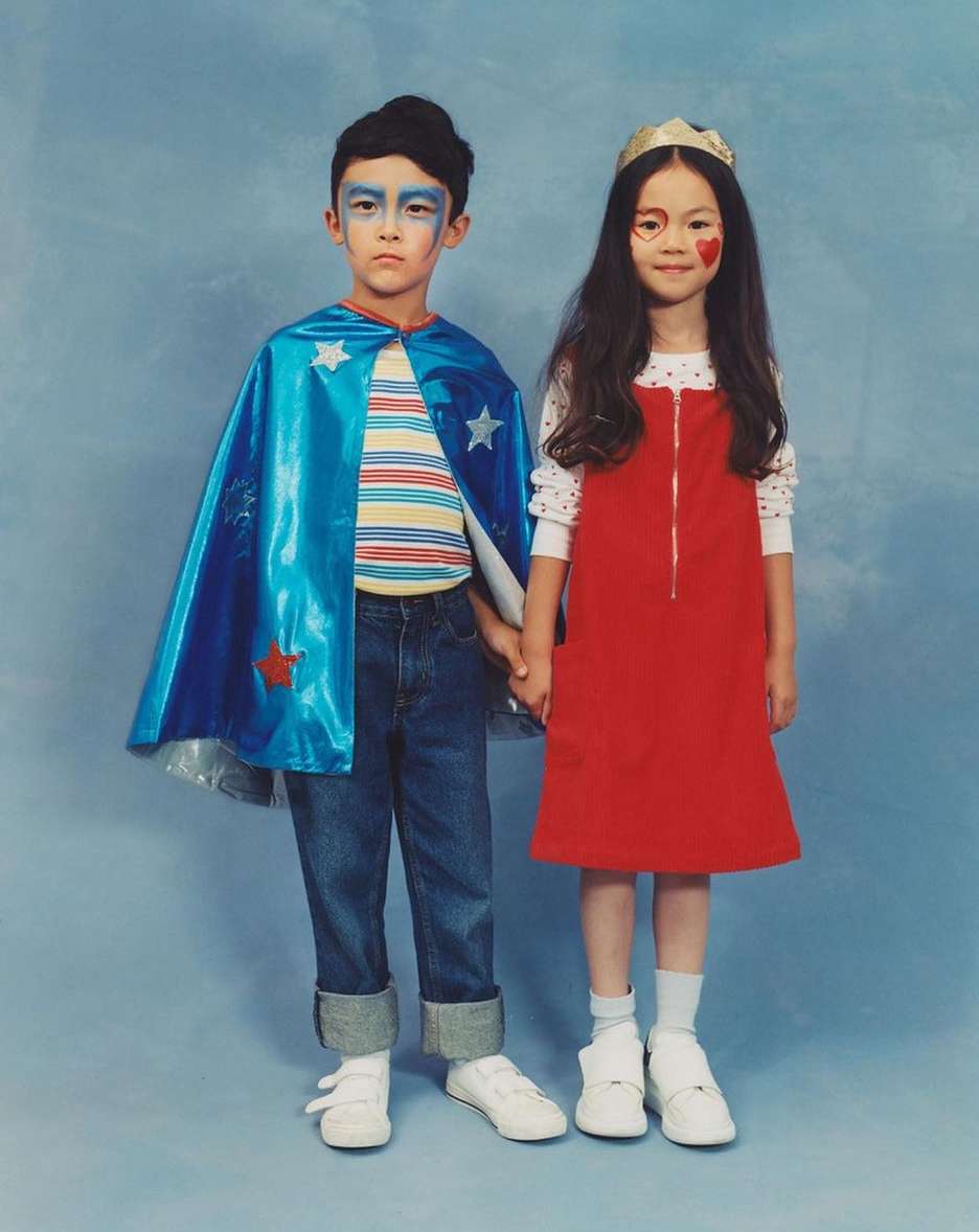 Two children dressed in costumes stand side-by-side against a mottled blue background, holding hands and looking forward.