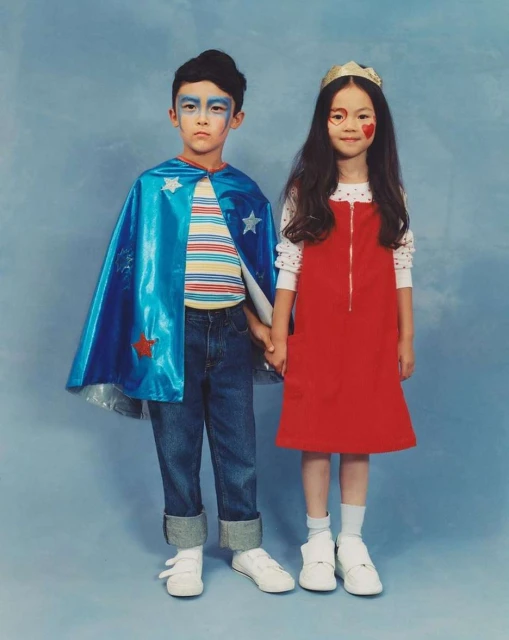 Two children dressed in costumes stand side-by-side against a mottled blue background, holding hands and looking forward.