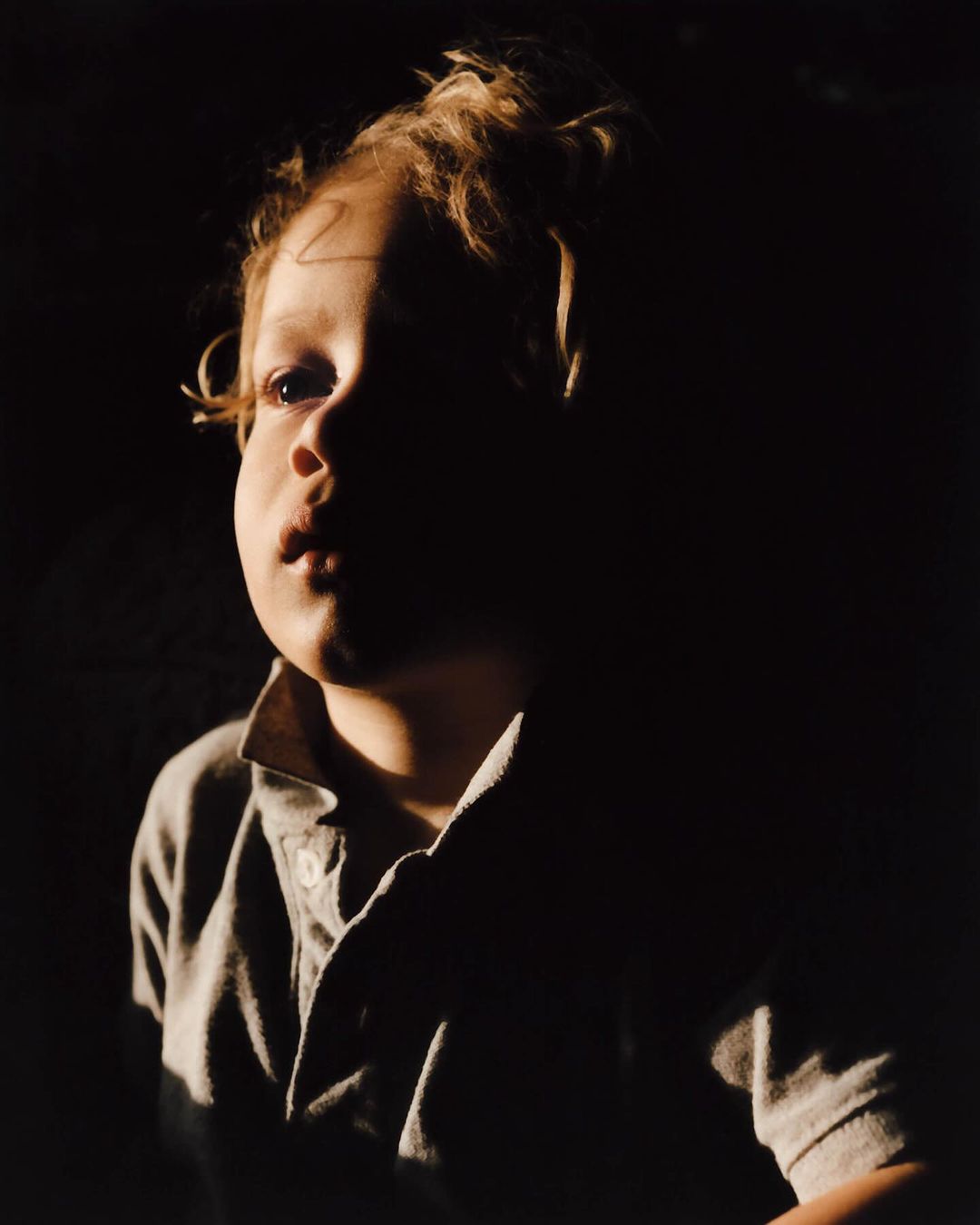 Portrait of a child with curly hair wearing a collared shirt under dramatic low-key lighting against a dark background