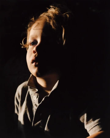 Portrait of a child with curly hair wearing a collared shirt under dramatic low-key lighting against a dark background