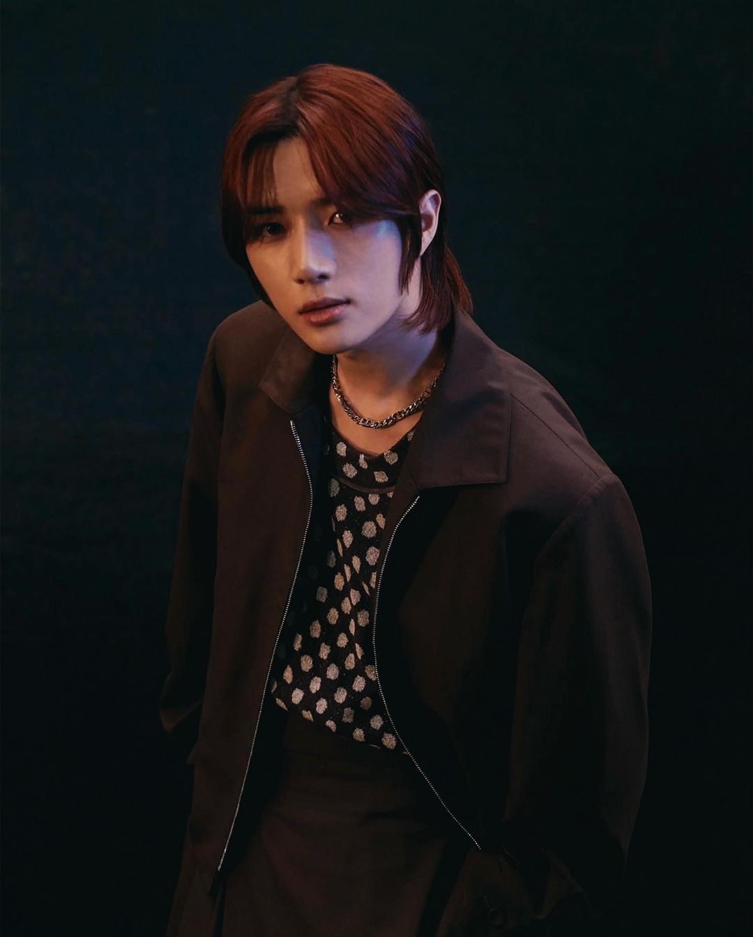 Portrait of a young adult male in a dark brown jacket over a polka dot shirt, posing in front of a dark background with dramatic lighting.