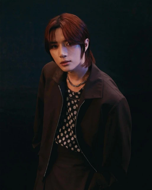 Portrait of a young adult male in a dark brown jacket over a polka dot shirt, posing in front of a dark background with dramatic lighting.