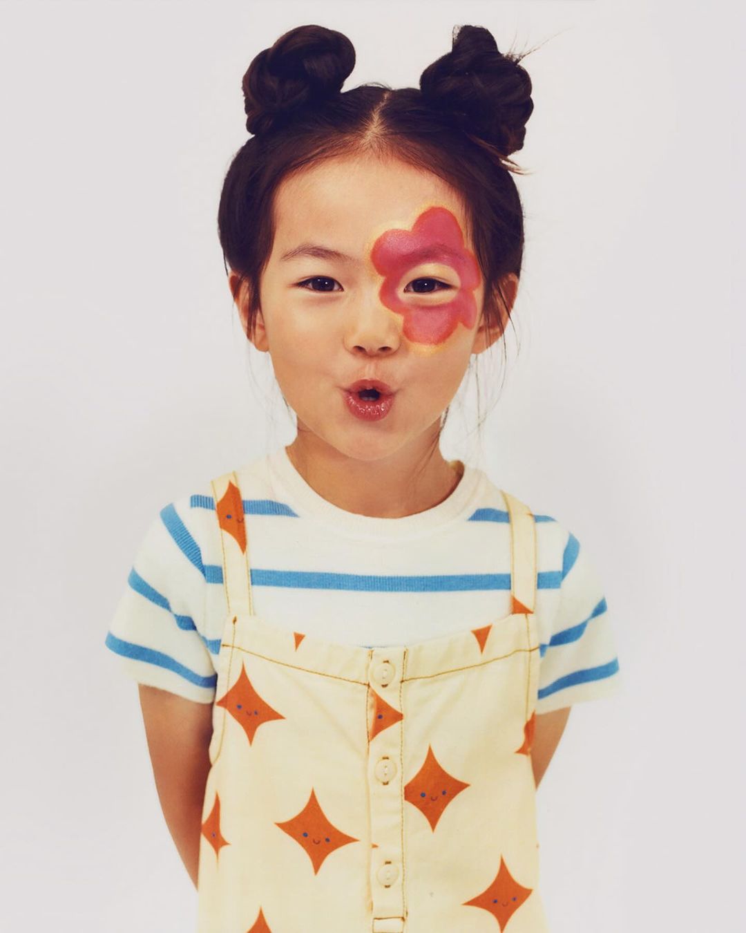Young girl with a flower painted on her face, wearing a striped shirt and patterned dress, posing in front of a white background.