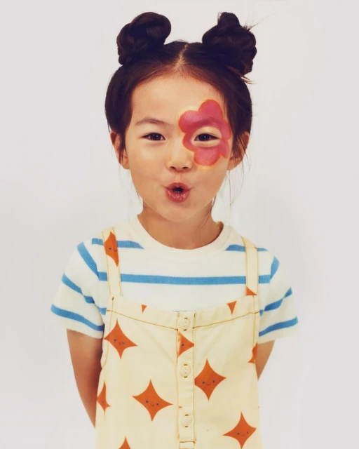 Young girl with a flower painted on her face, wearing a striped shirt and patterned dress, posing in front of a white background.
