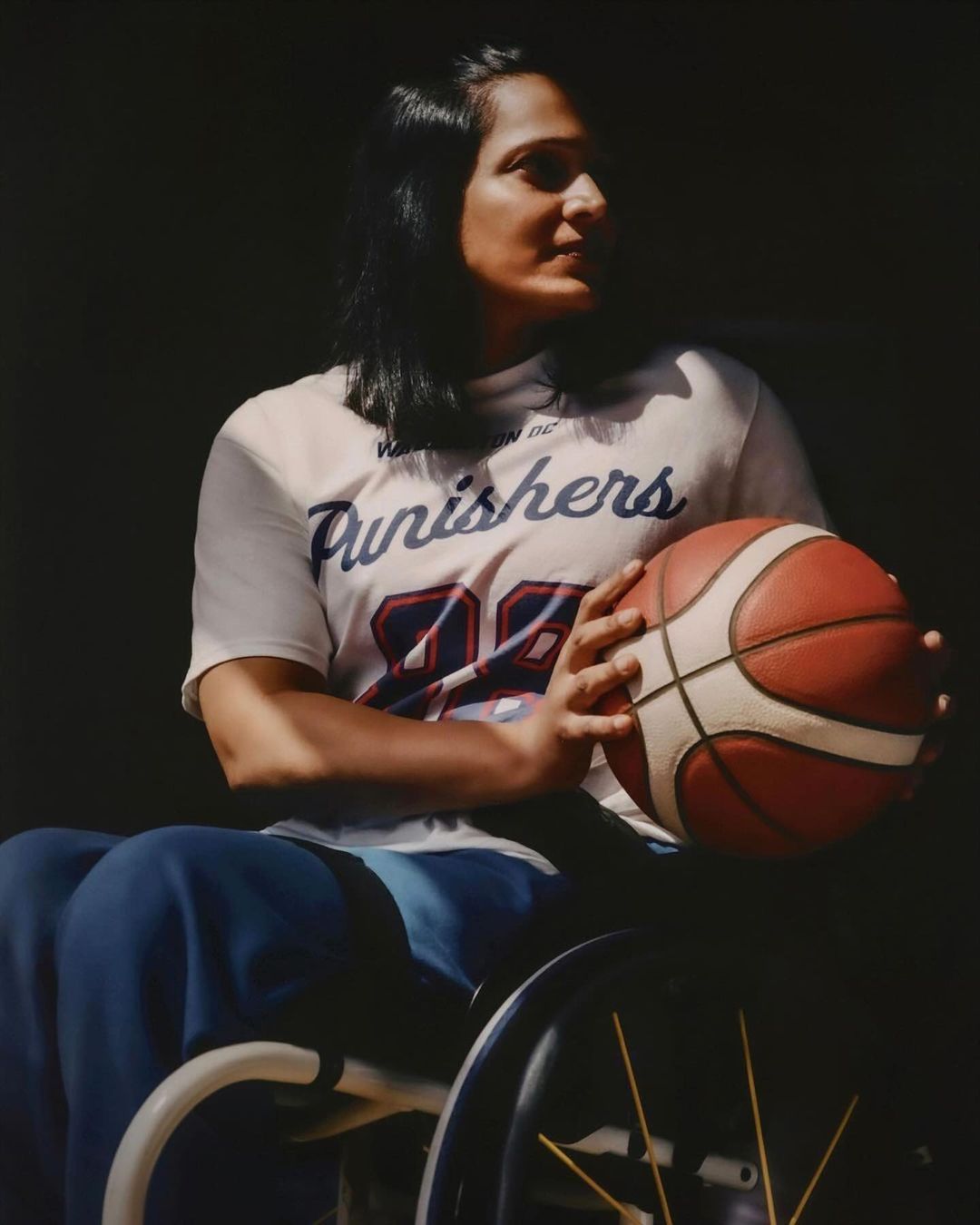 Athlete in wheelchair holds basketball with Punishers logo T-shirt in an indoor setting with dark background, eye-level shot.