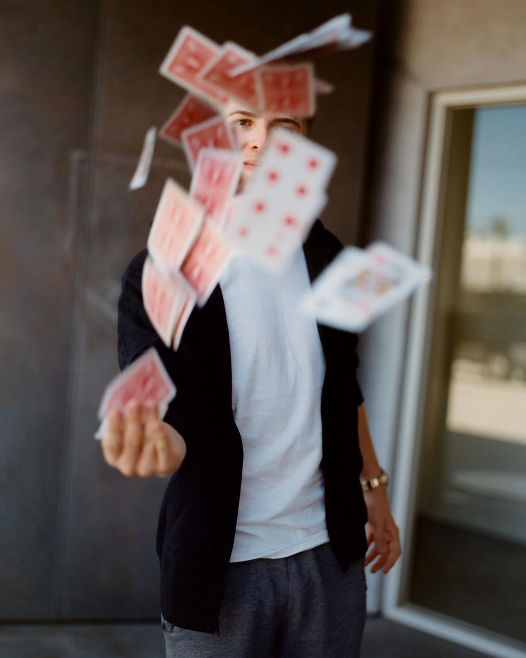 Young adult throwing a cascade of playing cards, wearing a white t-shirt and black cardigan in front of a modern grey building.