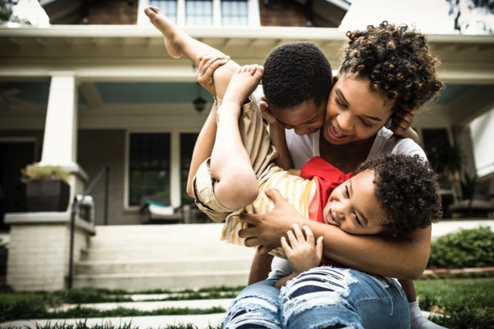 A joyous outdoor scene captures a Black mother embracing her two children, with a house visible in the background, under natural lighting.