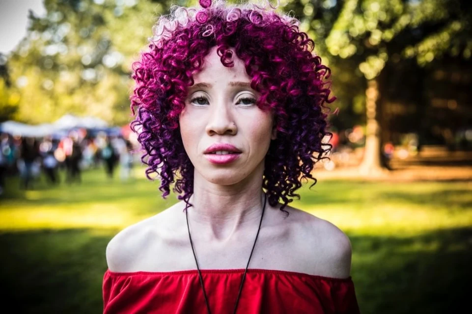 Close-up portrait of a woman with pink and purple curly hair, wearing a red off-the-shoulder top, standing in a grassy outdoor setting.