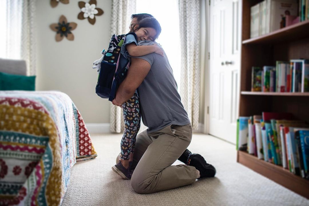 A father kneeling and hugging his young daughter with a backpack in a brightly lit home interior, showcasing a tender family moment.