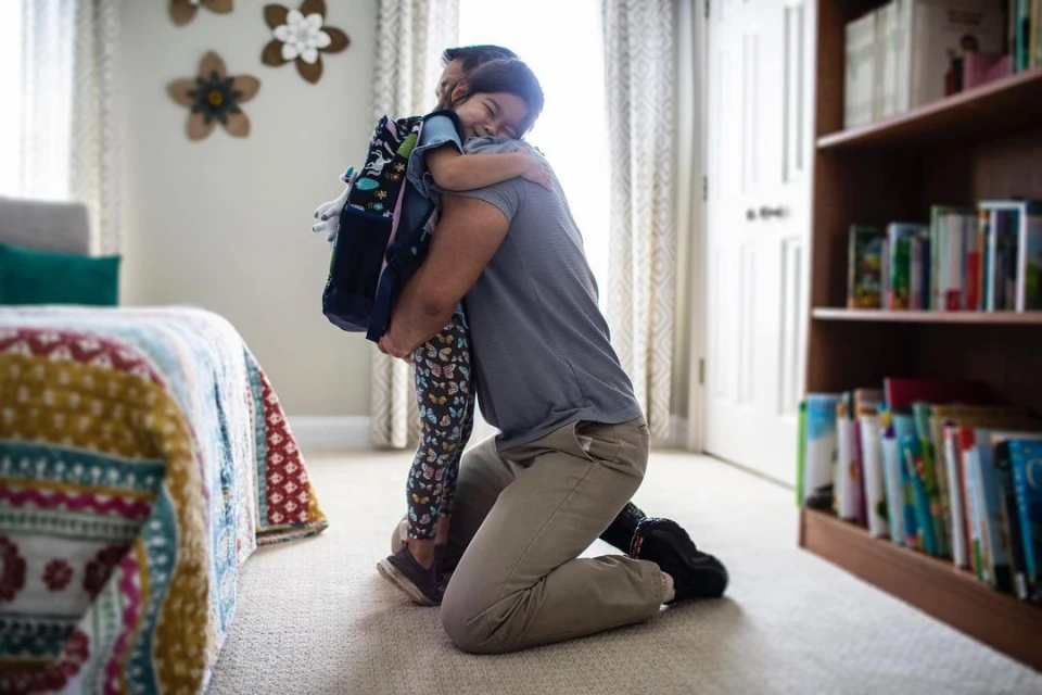 A father kneeling and hugging his young daughter with a backpack in a brightly lit home interior, showcasing a tender family moment.