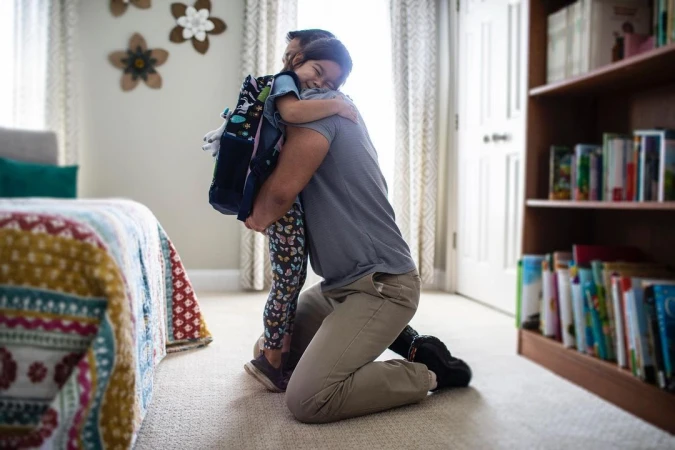 A father kneeling and hugging his young daughter with a backpack in a brightly lit home interior, showcasing a tender family moment.