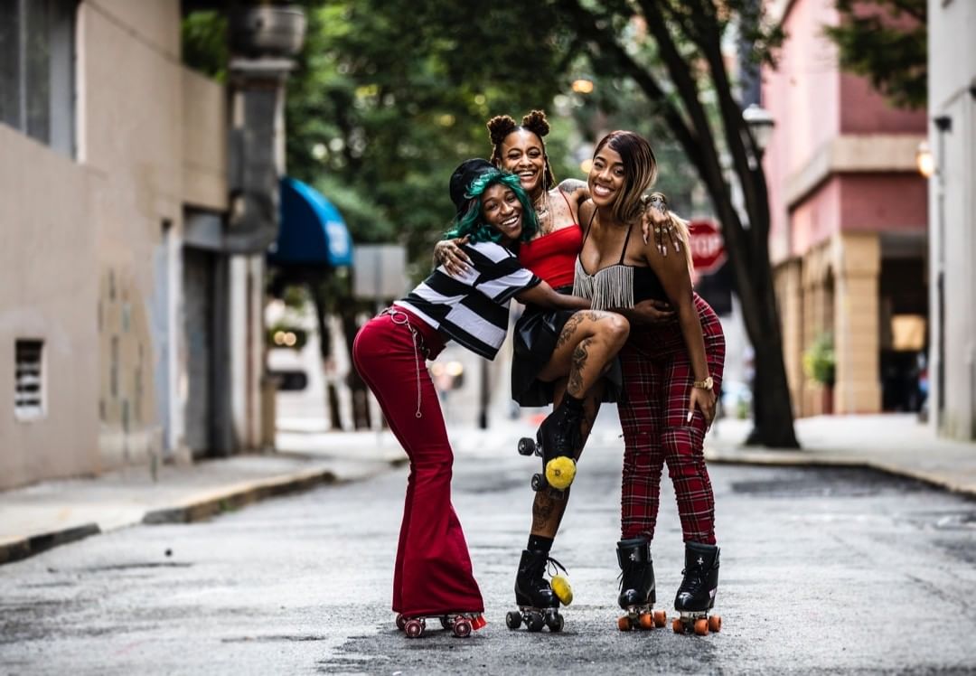 Three adult females pose on roller skates in an urban alleyway with brick buildings. They are smiling and wearing colorful clothing.
