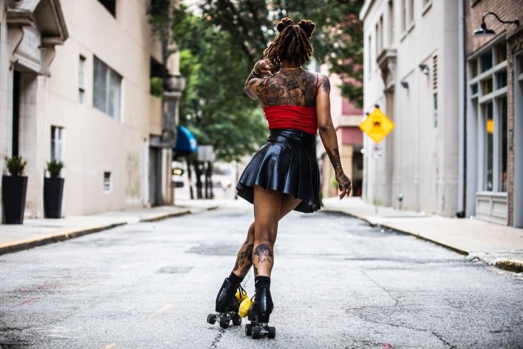Woman with tattoos skates on an empty street wearing a red top and black skirt. Buildings line the road, viewed from a low angle.