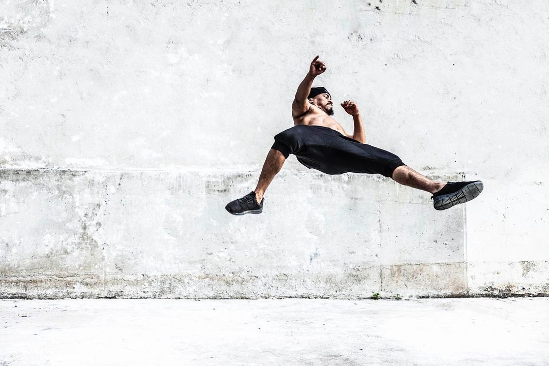 Athletic man in sportswear mid-air leaps against a textured concrete wall. Dark athletic wear and sneakers provide a striking silhouette.