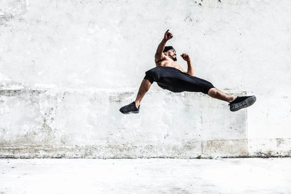 Athletic man in sportswear mid-air leaps against a textured concrete wall. Dark athletic wear and sneakers provide a striking silhouette.
