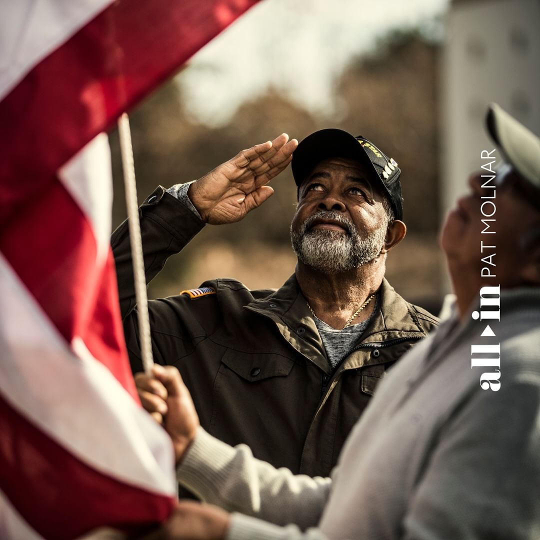 Man in military jacket salutes American flag held by another person outdoors, shot at eye level with medium framing in natural light.