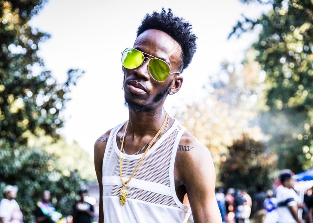 Portrait of an adult Black man wearing yellow-tinted aviator sunglasses and a white mesh tank top outdoors in a wooded area.