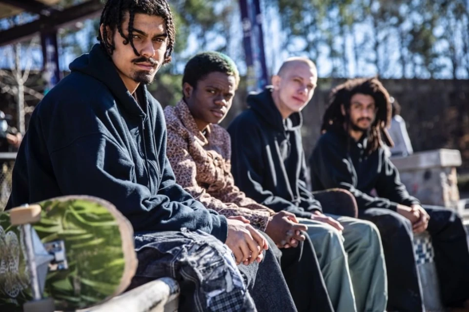 Group of young diverse men with skateboard outdoors. Four skateboarders sitting together outside in daylight, wearing casual streetwear.