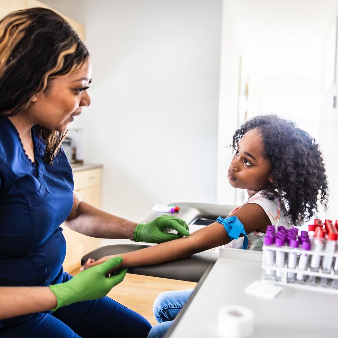 Healthcare worker in blue scrubs prepares a young patient for a blood draw, using a tourniquet and wearing green gloves in a clinic setting.