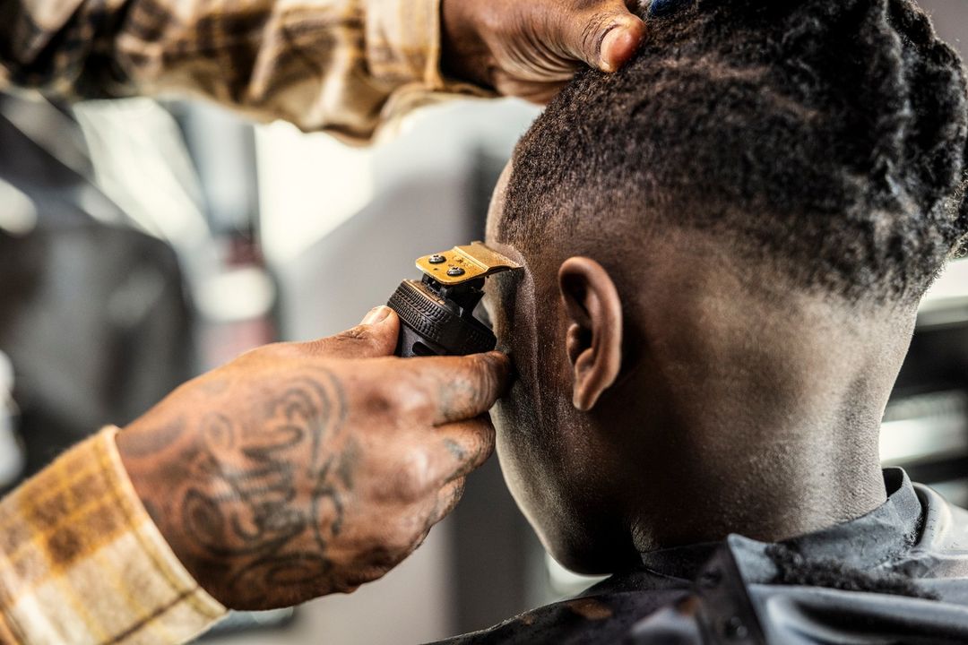 Barber giving a haircut to a young man with a hair clipper, close-up in an indoor setting. Man's hand is tattooed.