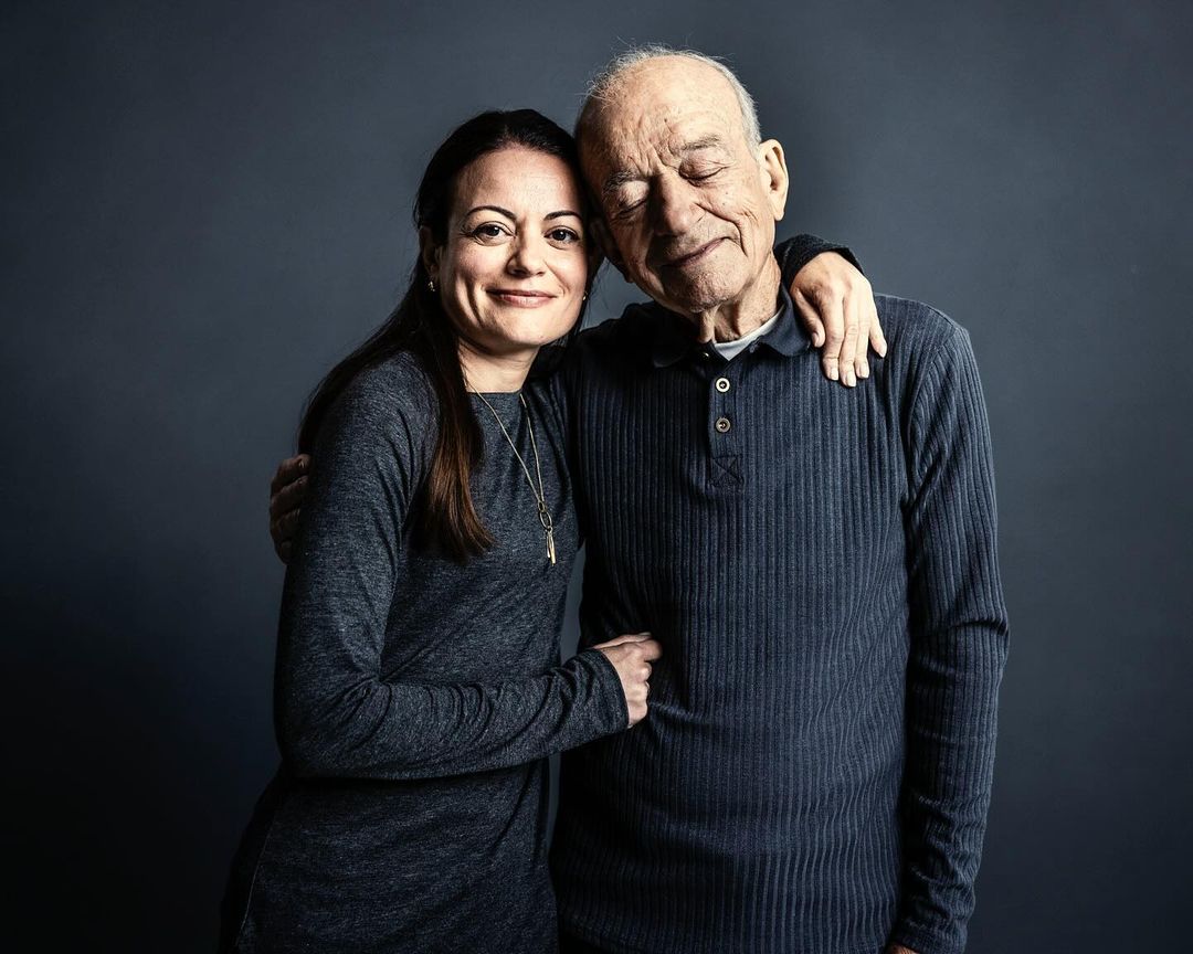 Portrait of a smiling adult woman embracing an elderly man in front of a dark gray background, showcasing a loving relationship between generations.