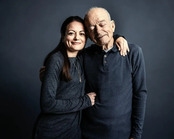Portrait of a smiling adult woman embracing an elderly man in front of a dark gray background, showcasing a loving relationship between generations.
