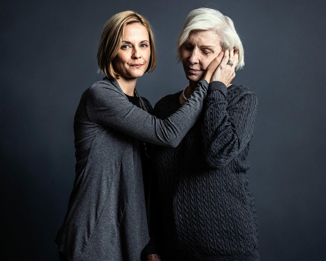 Portrait of a fair-skinned woman in a grey cardigan embracing an elderly fair-skinned woman in a grey sweater in front of a grey backdrop.