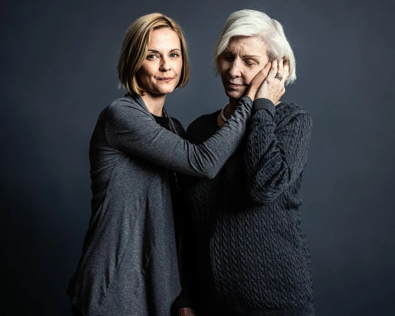 Portrait of a fair-skinned woman in a grey cardigan embracing an elderly fair-skinned woman in a grey sweater in front of a grey backdrop.