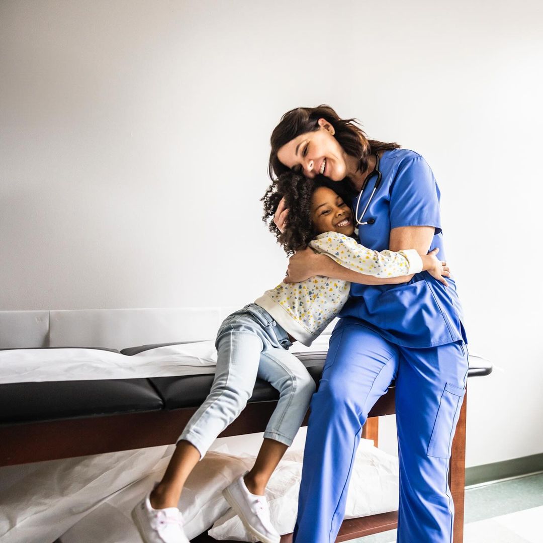 Healthcare scene shows a female nurse in blue scrubs hugging a smiling African American child in a doctor's office, fostering comfort.