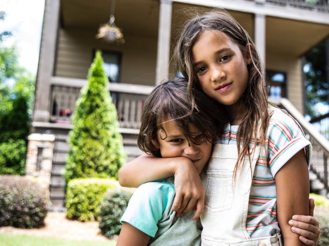 Two children, a girl and a boy, embrace in front of a house surrounded by greenery, with a soft, natural outdoor lighting.