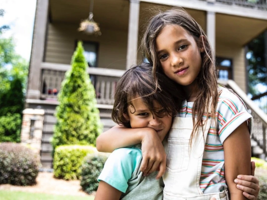 Two children, a girl and a boy, embrace in front of a house surrounded by greenery, with a soft, natural outdoor lighting.