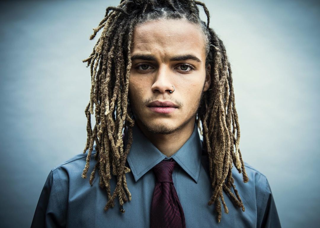 Portrait of a young adult male with dreadlocks, wearing a grey button-down shirt and a burgundy tie against a neutral background.