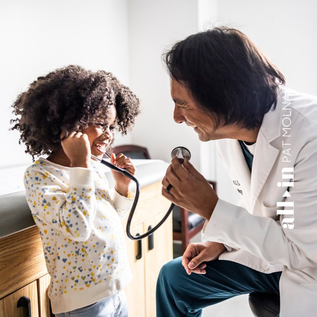 Child is examined by doctor in bright medical office. Girl holds stethoscope to her ears as the doctor looks on and smiles.
