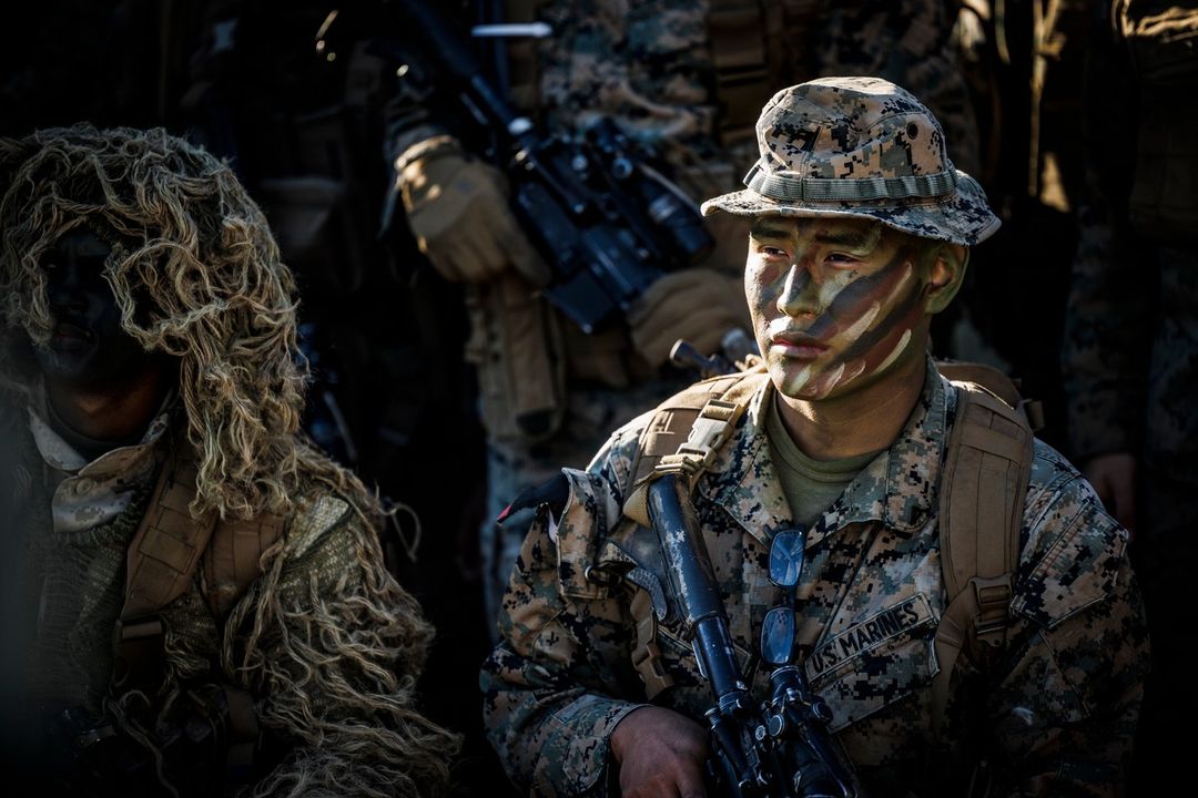 Camouflaged US Marines in military gear with rifles, wearing camo hats and ghillie suits, standing outdoors with natural lighting.