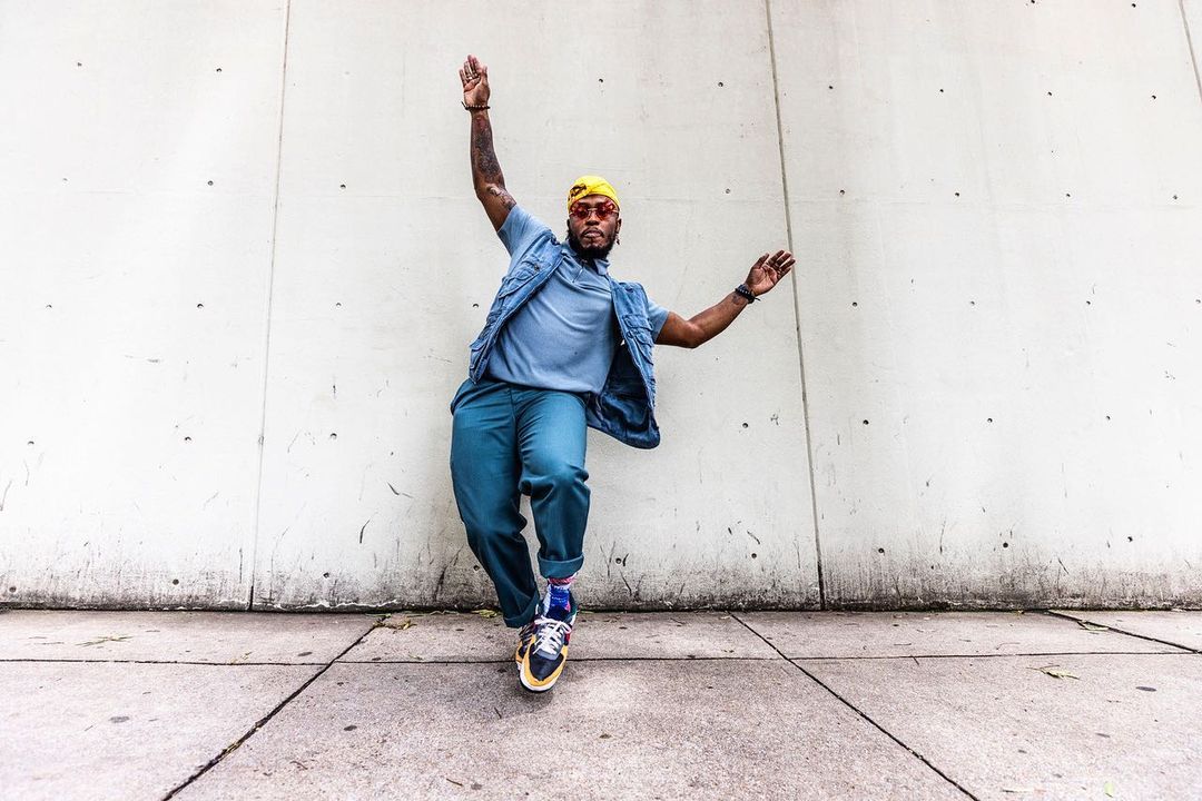 Stylish African-American man in casual wear dances against an urban concrete wall with denim vest, yellow bandana, and blue slacks.