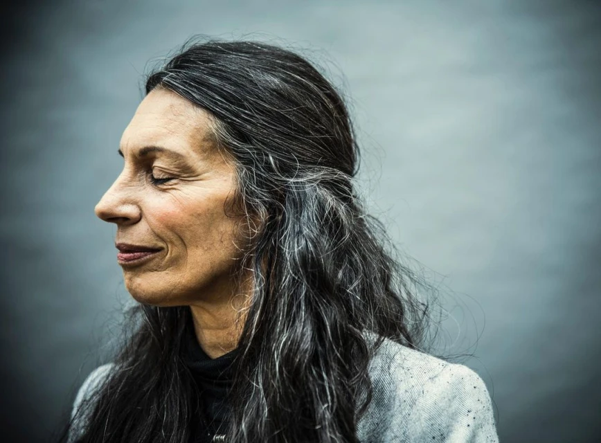 Portrait of a mature adult female in profile with eyes closed, wearing a grey jacket, against a muted blue-grey background in a close-up shot.