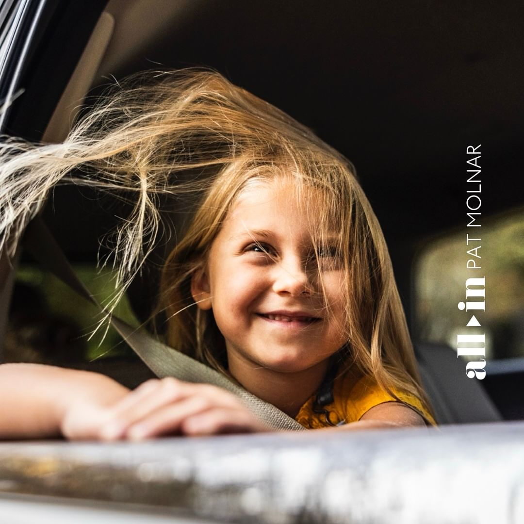 Close-up of a smiling child with blonde hair inside of a car, wearing a seatbelt, with text 'all in PAT MOLNAR' visible on the right.